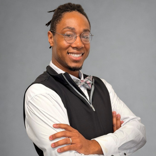 Smiling man with glasses, bow tie, and vest, arms crossed against a gray background.