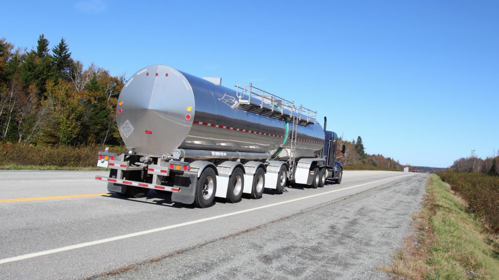 A polished silver tanker truck traveling on a rural two-lane highway with trees on the left and a clear blue sky.
