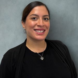 paasadena-legal-assistant Smiling woman with dark hair tied back, wearing a black top and jacket, heart pendant necklace, neutral gray background.